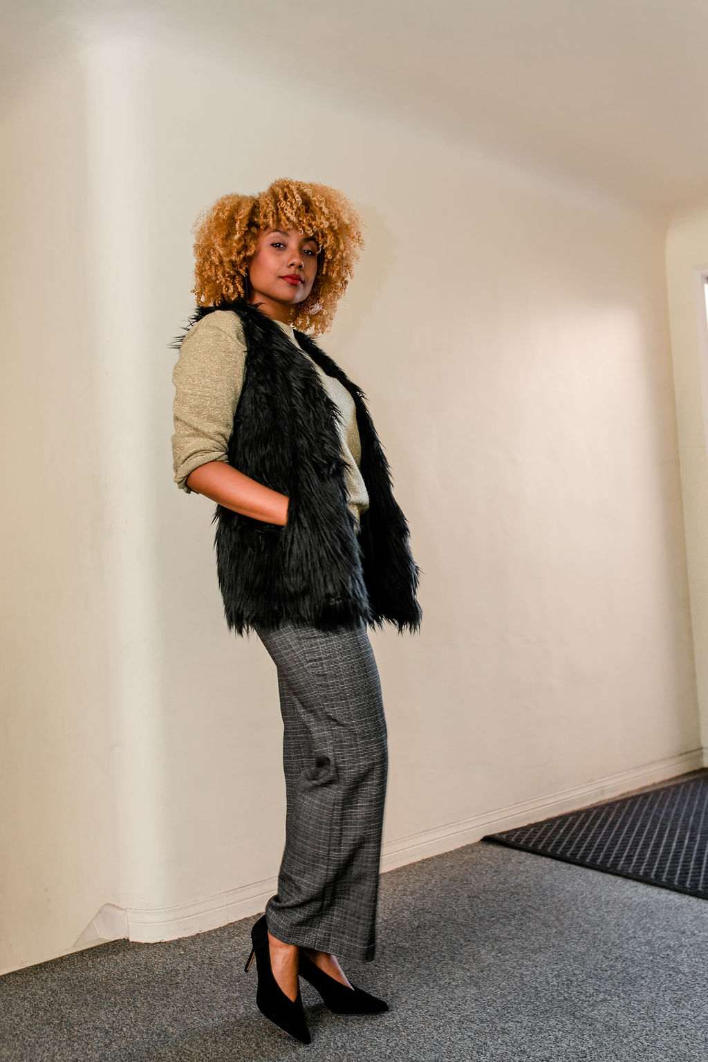 Black girl with curly blonde hair wearing slacks, fur vest and gold sweater- reflect